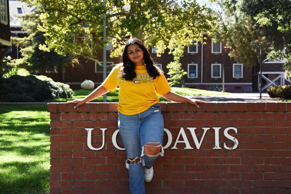 Aman Kaur leans against the red brick UC Davis sign.