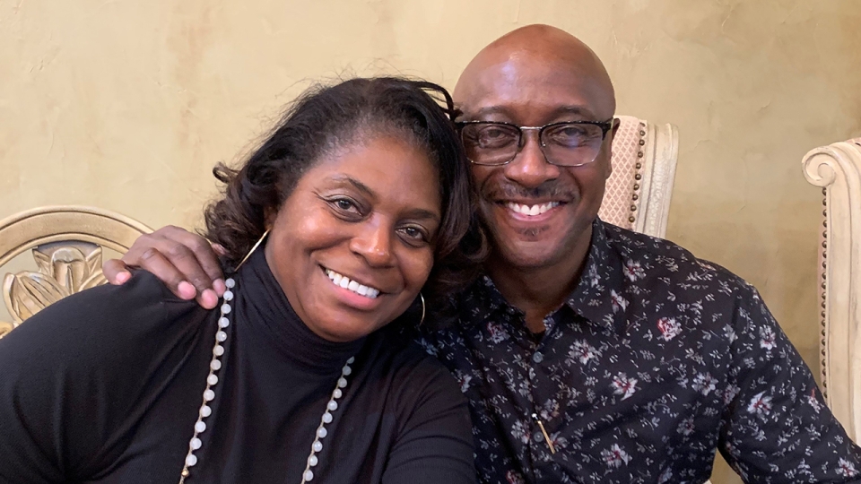 Lois wearing a black turtleneck leans against Darryl wearing glasses and a dark shirt with floral print.