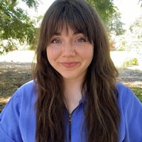 Portrait of Tessa Haden wearing a blue blouse in front sunlight casting a glow over trees and a walking path.